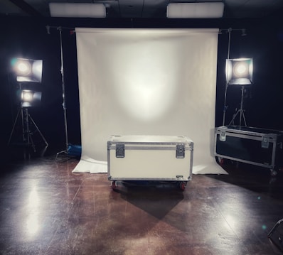 A photography studio setup featuring a large white backdrop, flanked by two professional studio lights on stands. A large, white equipment case with metal edges and handles is positioned in front of the backdrop. The floor is dark and glossy, reflecting some of the light.