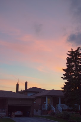 A suburban house is silhouetted against a vibrant dusk sky painted in shades of warm pink and orange. The foreground features a driveway leading up to a garage, flanked by a well-maintained yard and a large evergreen tree on the right.