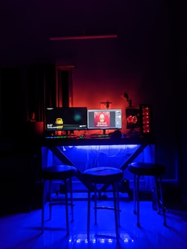 A dimly lit room features a modern computer setup on a desk with two monitors and a glowing CPU. The room is primarily illuminated with blue and red LED lights creating a vibrant atmosphere. Three stools are positioned in front of the desk, and various cables are visible under the desk.