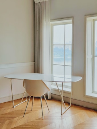 A minimalist interior scene featuring a white oval table with slender legs and a matching white chair. The room is well-lit by two tall windows with sheer curtains, allowing natural light to create a serene atmosphere. The herringbone pattern of the wooden floor adds a touch of warmth to the otherwise neutral-toned space.
