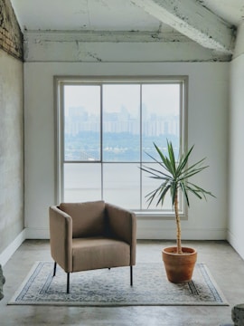 A minimalist interior features a single beige armchair on a patterned rug facing a large window. Outside, a distant view of a city skyline can be seen. A potted yucca plant stands beside the chair, complemented by a rough, industrial-style ceiling and walls.