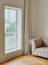 A room with a large window that has white curtains beside it. A tufted beige sofa with wooden legs is positioned near the window on a hardwood floor. Outside the window, there's a view of some trees and buildings in the background.