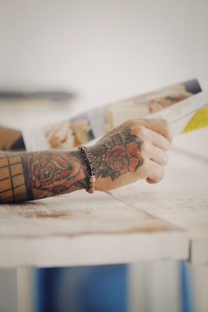 A tattooed hand holding a book or magazine, with the focus on the intricate floral tattoos and a beaded bracelet.
