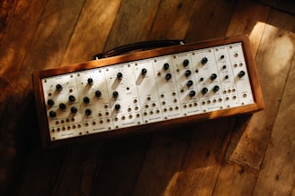 A modular synthesizer with multiple dials and switches mounted on a wooden frame. The synthesizer is resting on a rustic wooden floor with sunlight casting gentle shadows across its surface.