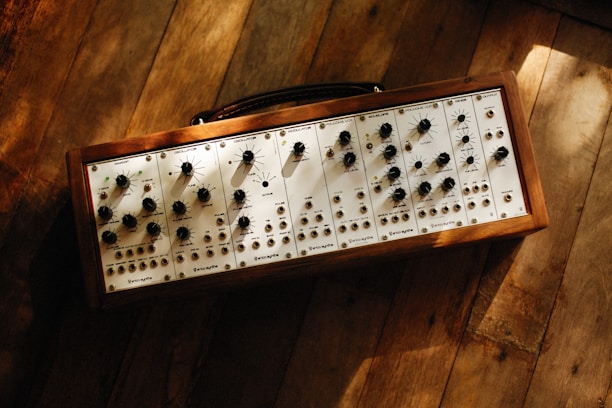 A modular synthesizer with multiple dials and switches mounted on a wooden frame. The synthesizer is resting on a rustic wooden floor with sunlight casting gentle shadows across its surface.