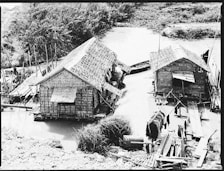 Two traditional thatched-roof houses are positioned on a riverbank with wooden pathways leading to them. The houses appear to be made from natural materials like bamboo and wood. Some boats are docked nearby, and there is a grassy area and dense vegetation in the background. Clothing is hung out to dry on lines, suggesting everyday life activities.