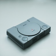 A classic Sony PlayStation console in gray, lying on a plain surface. The console features a disk drive in the center with buttons and controller ports on the front.