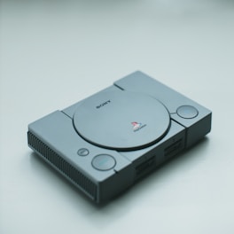 A classic Sony PlayStation console in gray, lying on a plain surface. The console features a disk drive in the center with buttons and controller ports on the front.
