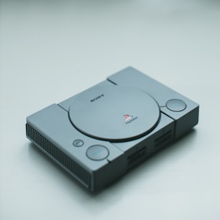 A classic Sony PlayStation console in gray, lying on a plain surface. The console features a disk drive in the center with buttons and controller ports on the front.