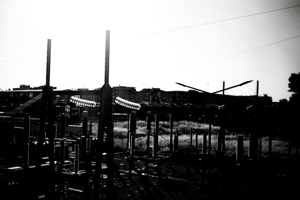 A black and white photograph of an industrial structure with tall pylons and metal frameworks. In the background, there are buildings set against a bright sky, creating strong contrasts and shadows. The geometric shapes of the machinery are prominent in the foreground.