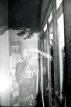 A black and white photograph depicts a person taking a picture with a camera. The scene is reflected in a window, creating a layered effect. The image includes reflections of trees and the interior, conveying a sense of depth and merging of indoor and outdoor environments.