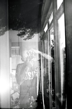 A black and white photograph depicts a person taking a picture with a camera. The scene is reflected in a window, creating a layered effect. The image includes reflections of trees and the interior, conveying a sense of depth and merging of indoor and outdoor environments.