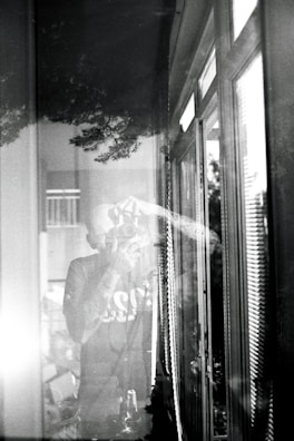 A black and white photograph depicts a person taking a picture with a camera. The scene is reflected in a window, creating a layered effect. The image includes reflections of trees and the interior, conveying a sense of depth and merging of indoor and outdoor environments.