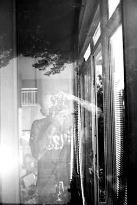 A black and white photograph depicts a person taking a picture with a camera. The scene is reflected in a window, creating a layered effect. The image includes reflections of trees and the interior, conveying a sense of depth and merging of indoor and outdoor environments.