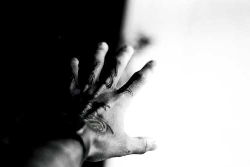 A black and white photograph featuring a hand with tattoos reaching towards a bright light source. The contrast between the dark and light areas creates a dramatic effect.