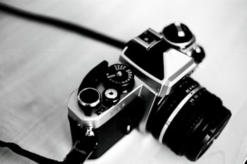 A vintage film camera with a classic design, featuring a large lens and numerous dials and switches on the top. The camera is angled towards the bottom left, set against a softly blurred background.