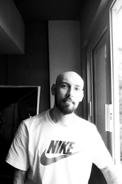 A person with a bald head and a beard is standing by a window, wearing a T-shirt with a recognizable sports brand logo. The image is in black and white, creating a contrast between light streaming in from the window and the shaded interior.