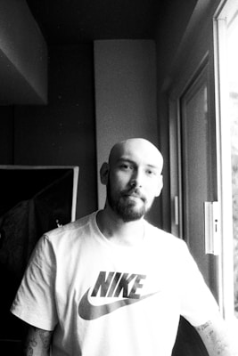 A person with a bald head and a beard is standing by a window, wearing a T-shirt with a recognizable sports brand logo. The image is in black and white, creating a contrast between light streaming in from the window and the shaded interior.