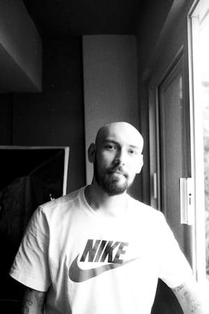 A person with a bald head and a beard is standing by a window, wearing a T-shirt with a recognizable sports brand logo. The image is in black and white, creating a contrast between light streaming in from the window and the shaded interior.