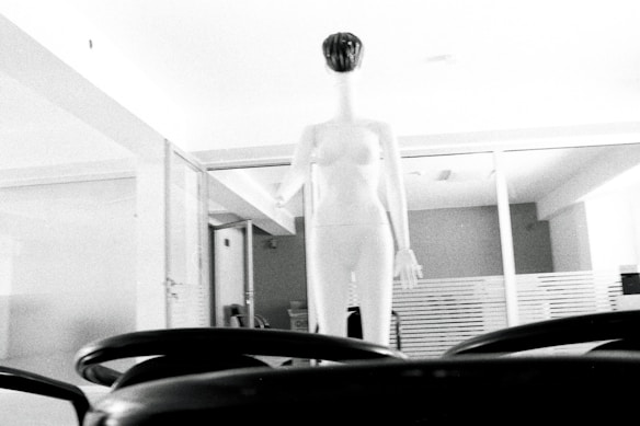 A minimalist interior space features a tall, white mannequin positioned prominently in the center, surrounded by glass partitions. The lighting is bright, casting an ethereal glow that highlights the mannequin's smooth, featureless surface. In the foreground, parts of a black office chair are visible, providing a contrast to the overall lightness of the scene.