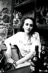 A person sitting in a casual pose at an indoor setting, wearing a t-shirt with printed graphics. Two bottles are placed in the foreground on a table. The surroundings include shelves with vintage fans and a stone wall, creating a slightly retro atmosphere.