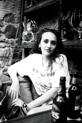 A person sitting in a casual pose at an indoor setting, wearing a t-shirt with printed graphics. Two bottles are placed in the foreground on a table. The surroundings include shelves with vintage fans and a stone wall, creating a slightly retro atmosphere.