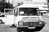A vintage-looking van parked in an urban setting is visible, with the front door open. The vehicle features signage in a foreign language, likely advertising products or services. Surrounding the van are a few parked cars and a building in the background, suggesting a city or town environment.