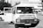 A vintage-looking van parked in an urban setting is visible, with the front door open. The vehicle features signage in a foreign language, likely advertising products or services. Surrounding the van are a few parked cars and a building in the background, suggesting a city or town environment.