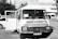 A vintage-looking van parked in an urban setting is visible, with the front door open. The vehicle features signage in a foreign language, likely advertising products or services. Surrounding the van are a few parked cars and a building in the background, suggesting a city or town environment.