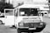 A vintage-looking van parked in an urban setting is visible, with the front door open. The vehicle features signage in a foreign language, likely advertising products or services. Surrounding the van are a few parked cars and a building in the background, suggesting a city or town environment.