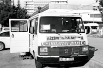 A vintage-looking van parked in an urban setting is visible, with the front door open. The vehicle features signage in a foreign language, likely advertising products or services. Surrounding the van are a few parked cars and a building in the background, suggesting a city or town environment.
