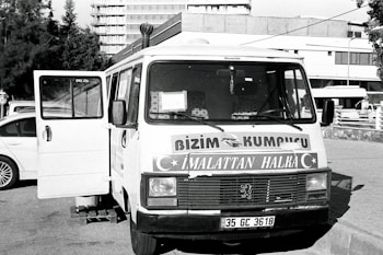 A vintage-looking van parked in an urban setting is visible, with the front door open. The vehicle features signage in a foreign language, likely advertising products or services. Surrounding the van are a few parked cars and a building in the background, suggesting a city or town environment.