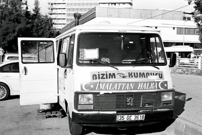 A vintage-looking van parked in an urban setting is visible, with the front door open. The vehicle features signage in a foreign language, likely advertising products or services. Surrounding the van are a few parked cars and a building in the background, suggesting a city or town environment.