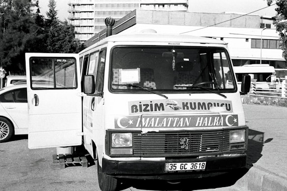 A vintage-looking van parked in an urban setting is visible, with the front door open. The vehicle features signage in a foreign language, likely advertising products or services. Surrounding the van are a few parked cars and a building in the background, suggesting a city or town environment.