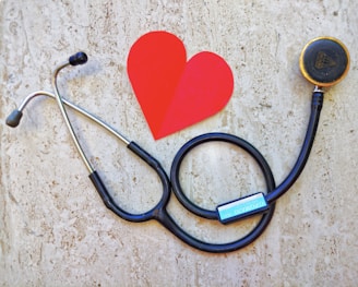 A stethoscope is arranged on a textured background in the shape of a heart next to a paper cutout of a red heart. The stethoscope's tubing forms a circular loop, with the chest piece near the red heart paper. The background has a stone-like appearance, adding contrast to the black tubing and bright red heart.