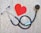 A stethoscope is arranged on a textured background in the shape of a heart next to a paper cutout of a red heart. The stethoscope's tubing forms a circular loop, with the chest piece near the red heart paper. The background has a stone-like appearance, adding contrast to the black tubing and bright red heart.
