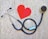 A stethoscope is arranged on a textured background in the shape of a heart next to a paper cutout of a red heart. The stethoscope's tubing forms a circular loop, with the chest piece near the red heart paper. The background has a stone-like appearance, adding contrast to the black tubing and bright red heart.