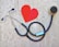 A stethoscope is arranged on a textured background in the shape of a heart next to a paper cutout of a red heart. The stethoscope's tubing forms a circular loop, with the chest piece near the red heart paper. The background has a stone-like appearance, adding contrast to the black tubing and bright red heart.