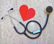 A stethoscope is arranged on a textured background in the shape of a heart next to a paper cutout of a red heart. The stethoscope's tubing forms a circular loop, with the chest piece near the red heart paper. The background has a stone-like appearance, adding contrast to the black tubing and bright red heart.