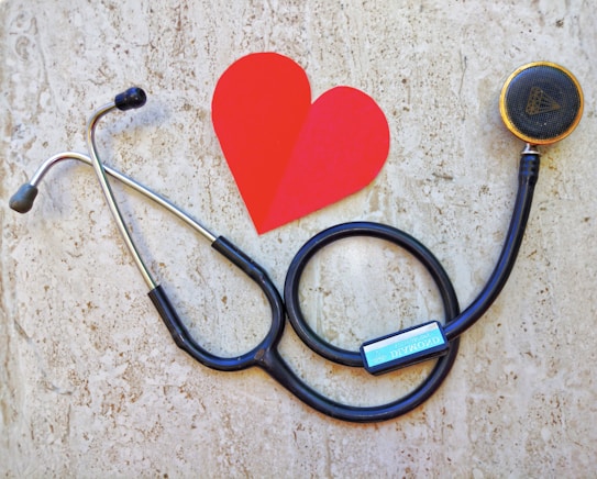 A stethoscope is arranged on a textured background in the shape of a heart next to a paper cutout of a red heart. The stethoscope's tubing forms a circular loop, with the chest piece near the red heart paper. The background has a stone-like appearance, adding contrast to the black tubing and bright red heart.