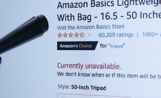 A webpage displaying a product listing for an Amazon Basics Lightweight Tripod. The product is currently unavailable, as indicated in red text. The listing includes a customer rating of five stars with 60,209 ratings, and it is marked as 'Amazon's Choice' for tripods. A black object, possibly part of the tripod, is visible in the foreground.