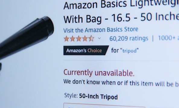 A webpage displaying a product listing for an Amazon Basics Lightweight Tripod. The product is currently unavailable, as indicated in red text. The listing includes a customer rating of five stars with 60,209 ratings, and it is marked as 'Amazon's Choice' for tripods. A black object, possibly part of the tripod, is visible in the foreground.