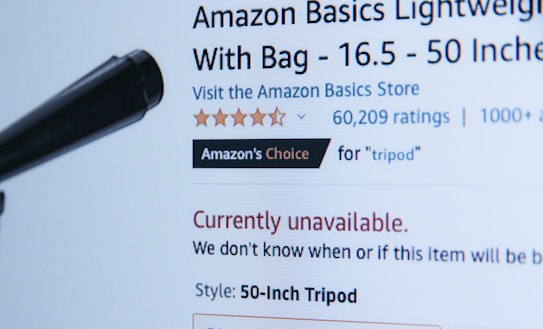 A webpage displaying a product listing for an Amazon Basics Lightweight Tripod. The product is currently unavailable, as indicated in red text. The listing includes a customer rating of five stars with 60,209 ratings, and it is marked as 'Amazon's Choice' for tripods. A black object, possibly part of the tripod, is visible in the foreground.