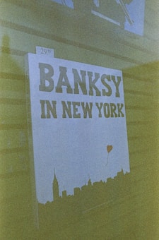 A canvas or poster displays the text 'Banksy in New York' prominently. Below the text is a silhouette of a city skyline, with what appears to be a small red balloon shaped like a heart floating above it. The price tag on the corner reads '$29.99'.