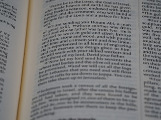A close-up view of an open book page displaying text in English. The layout appears to be similar to that of a religious or classical text, possibly a Bible, as indicated by the column format and verse numbers. The text discusses a message regarding a skilled individual named Huram-Abi and refers to craftsmanship in various materials.