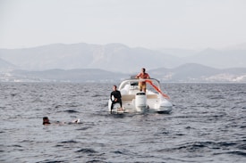 A motorboat is on a calm sea with three people present. One person is standing on the boat holding an orange flag, another is seated at the back of the boat, and the third person is swimming nearby. The background features distant mountains under a clear sky.