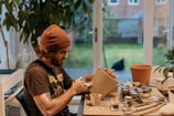 A man wearing a brown beanie and a dark t-shirt is sitting at a table inside a room with large windows facing a garden. He is painting a clay pot with numerous small paint containers and brushes scattered on the table. There are other clay pots and a plant nearby.