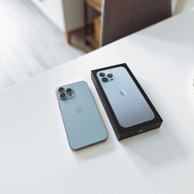 A smartphone is placed on a white surface next to its box. The phone and the box display a sleek, modern design with a metallic finish. The camera arrangement features multiple lenses, indicating advanced photographic capabilities.