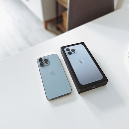 A smartphone is placed on a white surface next to its box. The phone and the box display a sleek, modern design with a metallic finish. The camera arrangement features multiple lenses, indicating advanced photographic capabilities.