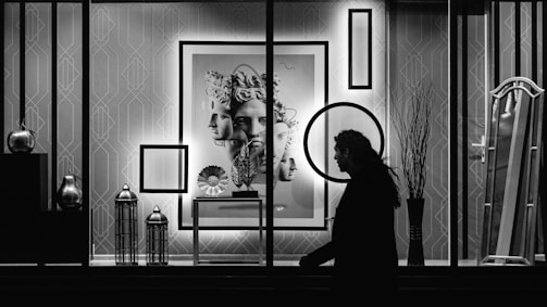 A black and white photo of an artful shop window display. The background features geometric patterned wallpaper, adorned with frames and a large artwork of a classical sculpture’s multiple faces. Various objects, such as vases and lanterns, are arranged on shelves. A silhouette of a person walking by is visible in the foreground, adding depth and contrast to the composition.